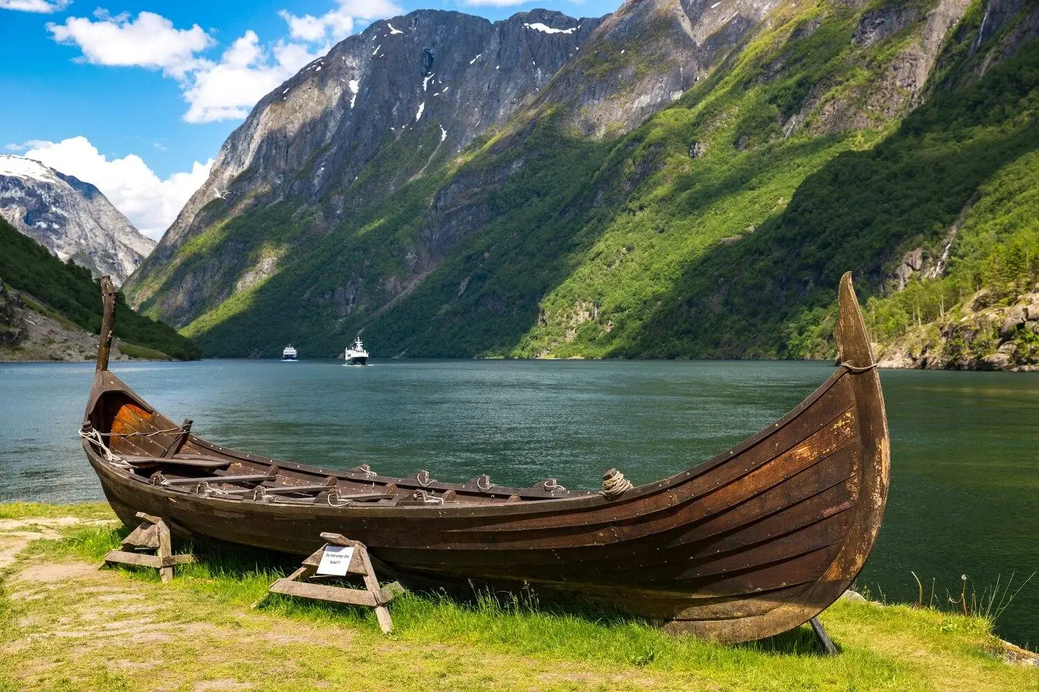 Ein hölzernes Wikingerschiff liegt am grasbewachsenen Ufer eines ruhigen Fjords, umgeben von steilen grünen Bergen und felsigen Klippen.