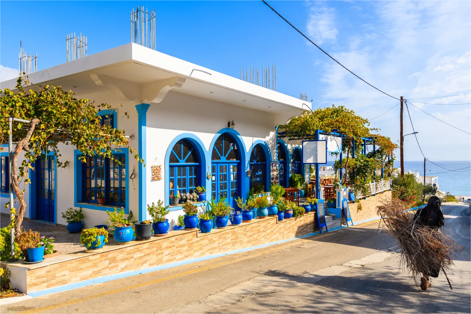 Alte Frau in schwarzem Kleid trägt Reisig und geht an einer Taverne vorbei auf der Straße in Diafani, Karpathos