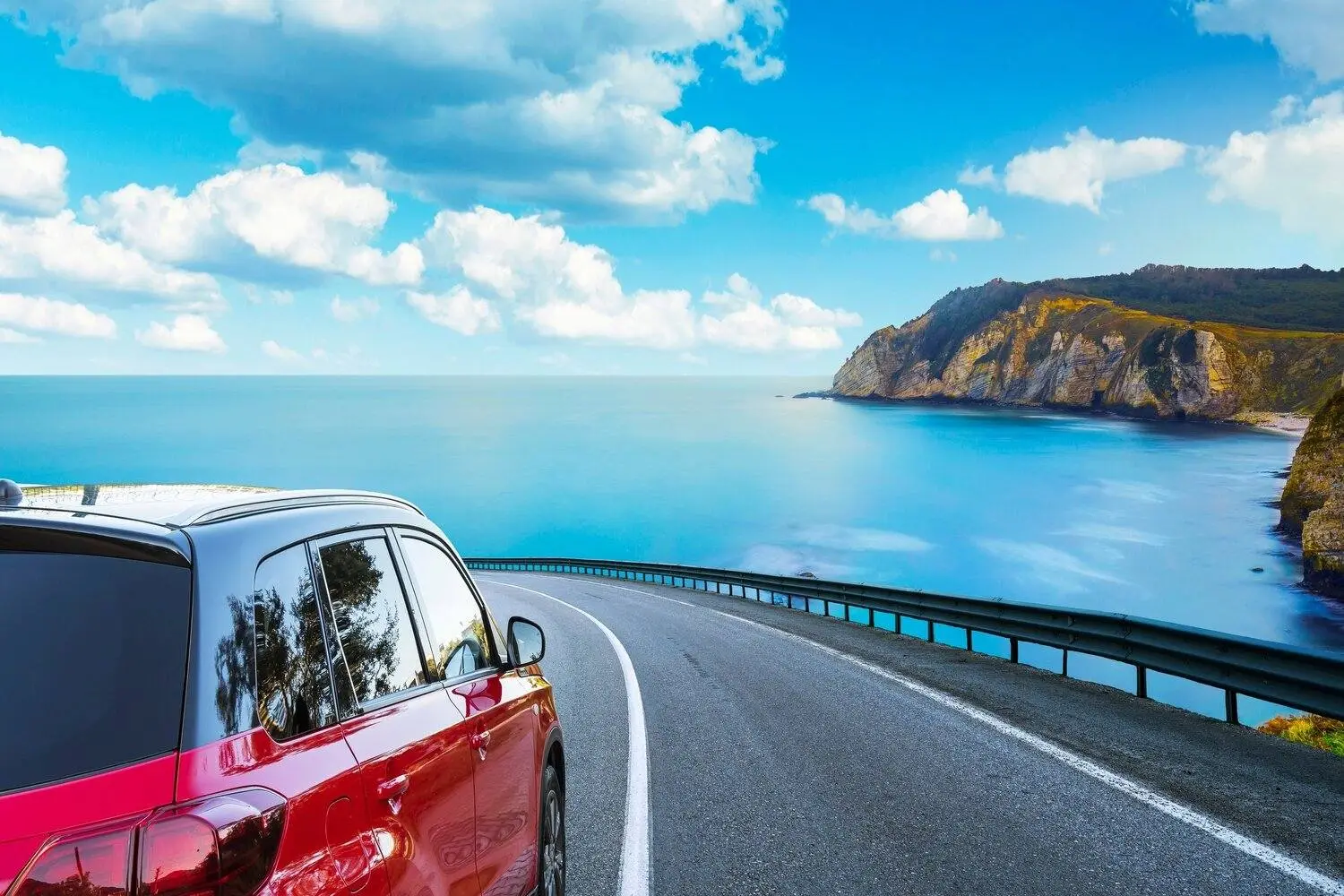 Rotes Auto fährt auf einer Küstenstraße mit Blick auf das blaue Meer und die felsigen Klippen unter einem hellen Himmel mit Wolken.